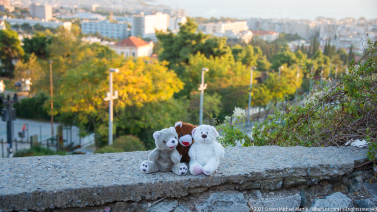 Three plush animals sitting on a rough stone wall above a leafy urban slope.