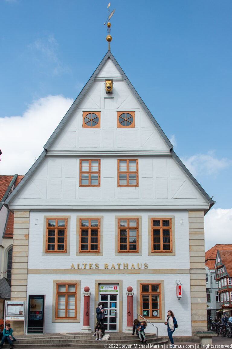 Pale gabled facade of Celle’s Altes Rathaus with orange-trimmed windows, central entrance, crest, weather vane, people, and bicycles.