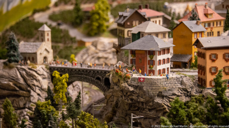 Stone arch bridge in miniature Swiss alpine village with pedestrians, church, colorful houses, rocky slopes, and Swiss flag.