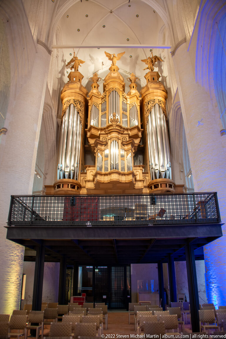 Large gilded wooden pipe organ with multiple pipe towers above a dark gallery in a white-brick vaulted church.