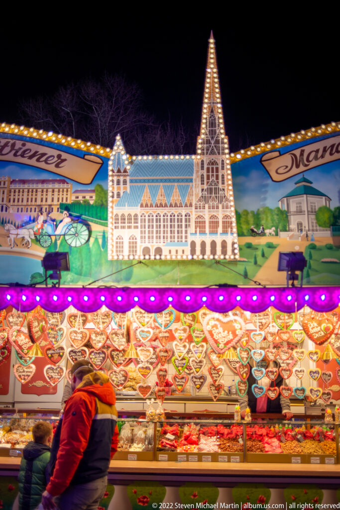 Sweet stall with rows of decorated treats beneath an illuminated Gothic-style church façade and painted cityscape panels.