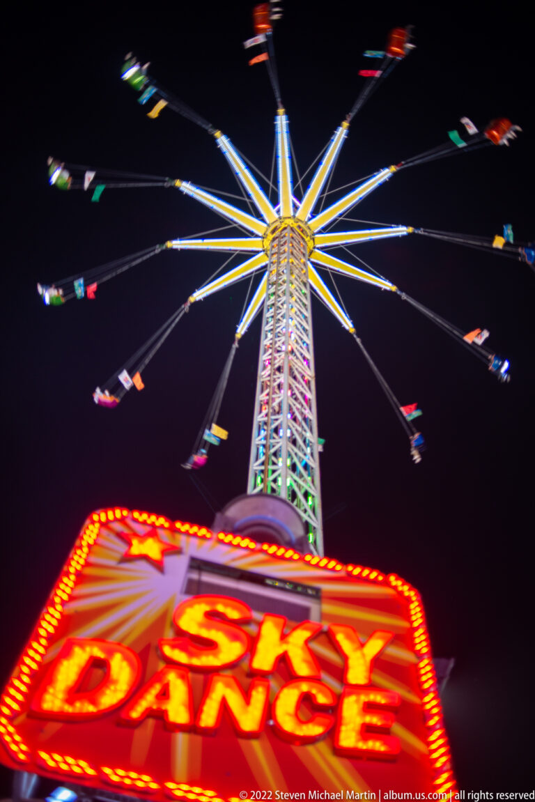 Red Sky Dance sign in the foreground with an illuminated tower swing ride spinning overhead at night.