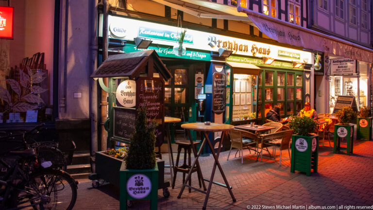 Illuminated Broyhan Haus pub and restaurant entrance with outdoor tables, signs, planters, and bicycles at dusk.