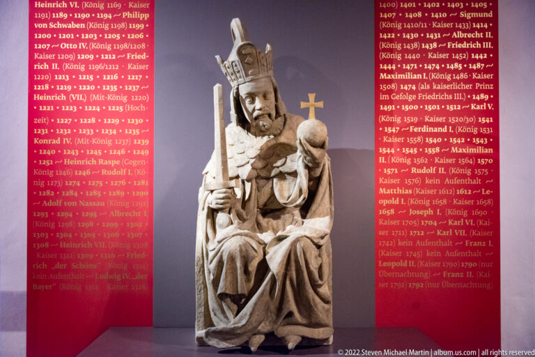 Seated sculpture of Emperor Heinrich VI wearing a crown and holding an orb and scepter before a red chronology panel.