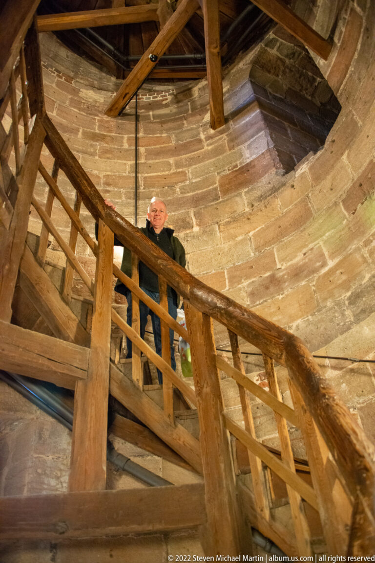 Visitor standing on steep wooden stairs and timber framing inside thick curved stone tower walls.