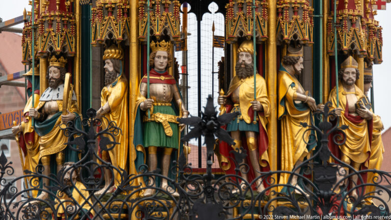 Close view of painted standing figures in Gothic niches on the Schöner Brunnen behind a black iron grille.