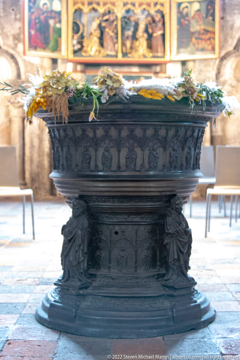 Large dark bronze baptismal font with relief figures around the basin and flowers resting on its rim.