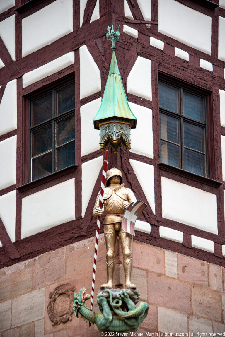 Small armored figure beneath a green copper canopy mounted against a red-and-white half-timbered façade.