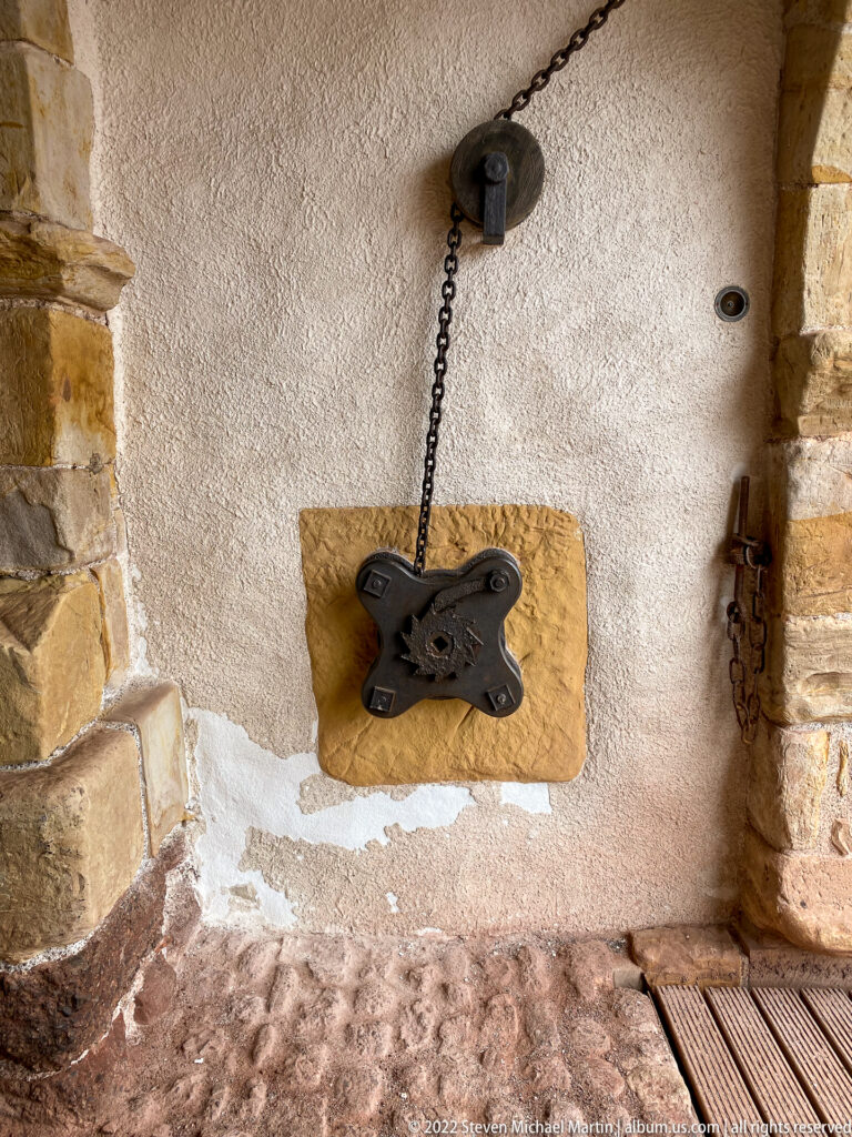 Black iron wall fitting mounted on wood and connected by a chain to a small overhead pulley in a plastered stone alcove.