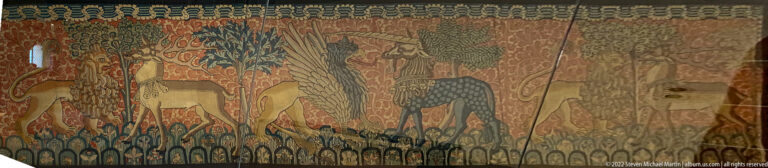 Historic tapestry with stylized hoofed animals, trees, dense foliage, and repeated decorative borders.