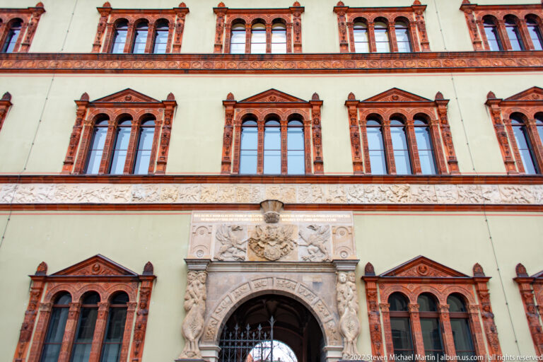 Central carved portal beneath symmetrical rows of red-framed windows on a pale historic façade.