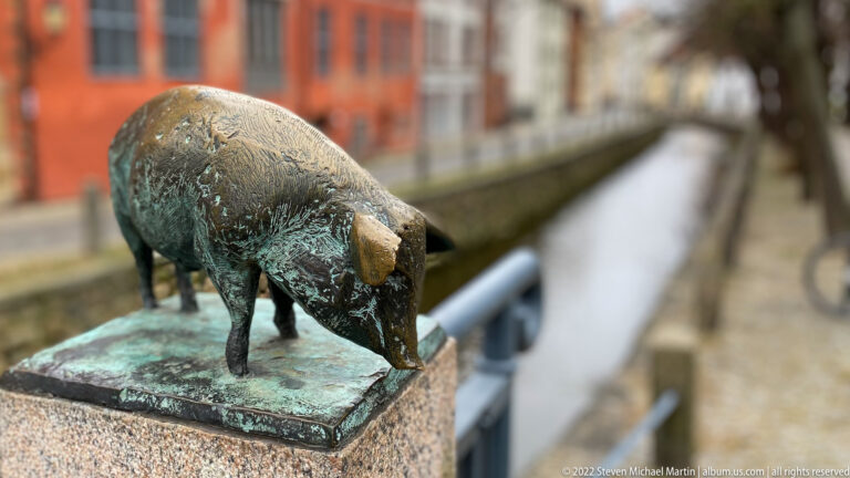 Standing bronze pig with worn snout and green patina on a post above a narrow canal.
