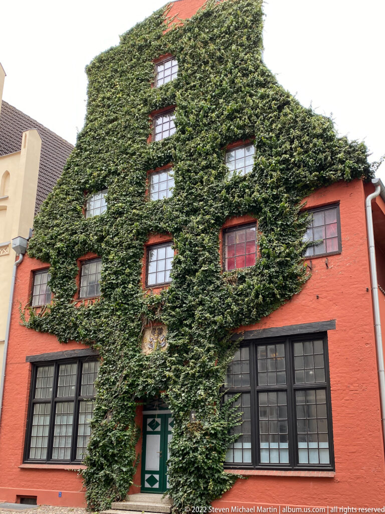 Dense ivy covers a red building with black-framed windows and a green-and-white doorway.