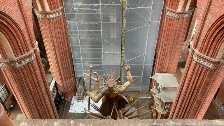 High view between red-brick church piers toward scaffolding, work platforms, and a large suspended sculptural figure.