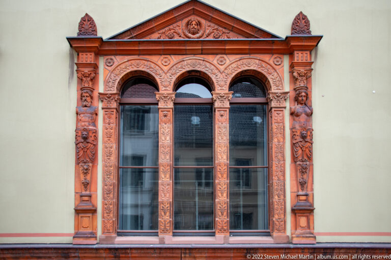 Three-part arched window with elaborate red carved surround, pilasters, masks, and triangular pediment.