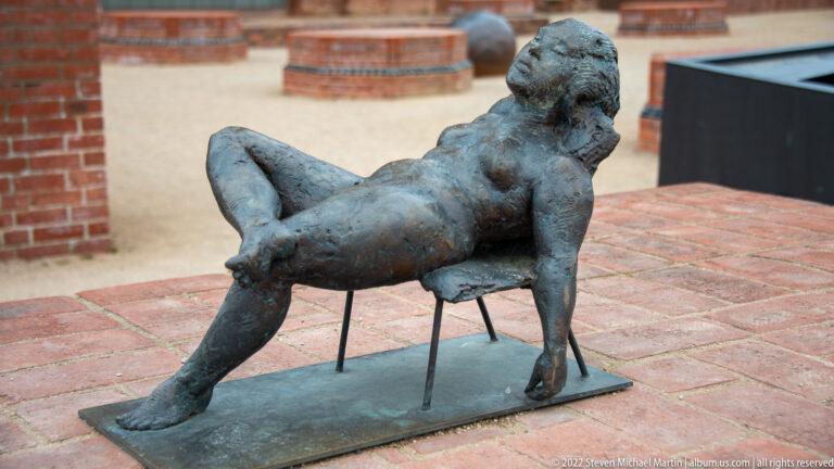 Small bronze sculpture of a reclining human figure on a low base beside brick paving.