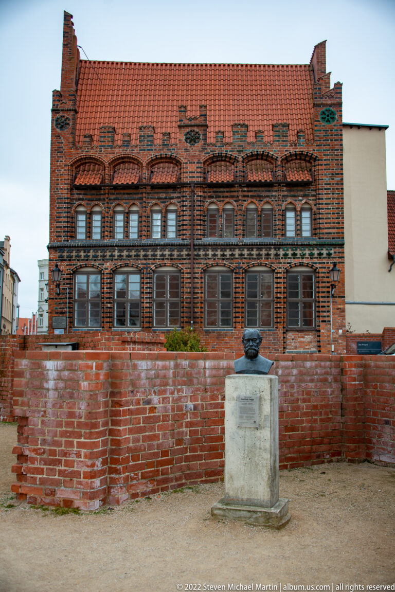 Decorated brick building with red tile roof behind a small bust monument and low brick wall.
