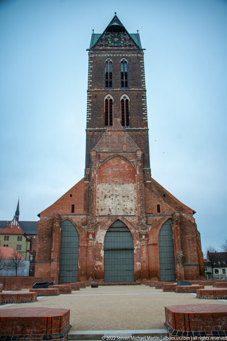 Massive square brick tower above truncated church walls with three large closed arches at ground level.