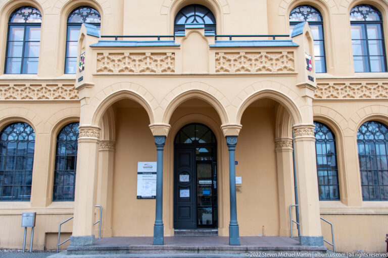 Triple pointed-arch entrance with slender columns, decorative tracery, shields, and tall flanking windows.