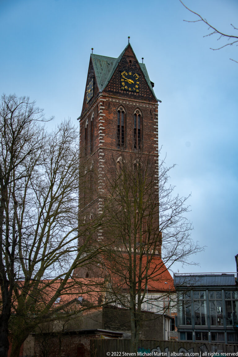 Square red-brick church tower with clock face, tall paired openings, and bare trees in front.