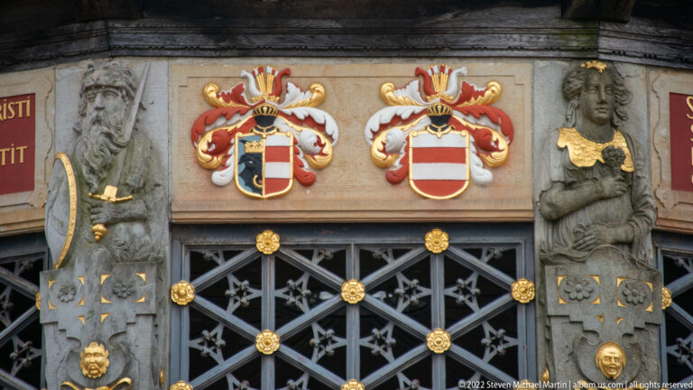 Two heraldic shields, sculpted figures, and a dark grille with gilded ornaments on the Wasserkunst.