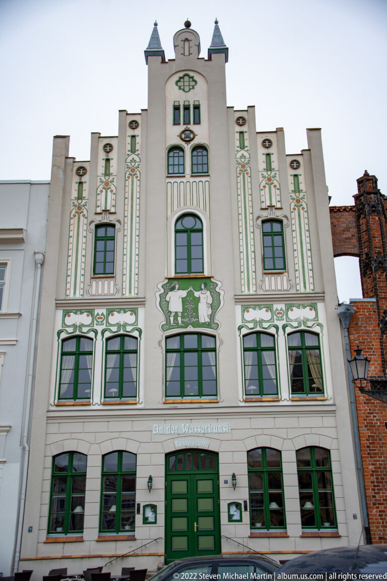 Tall white gabled façade with green windows, painted ornament, and symmetrical figurative panels.