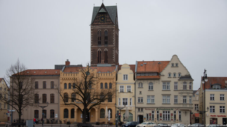 Tall brick church tower rising behind pale historic buildings and bare trees beside an urban square.
