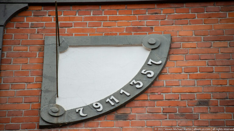 Semicircular sundial with numbered metal rim and projecting gnomon mounted on a red-brick wall.