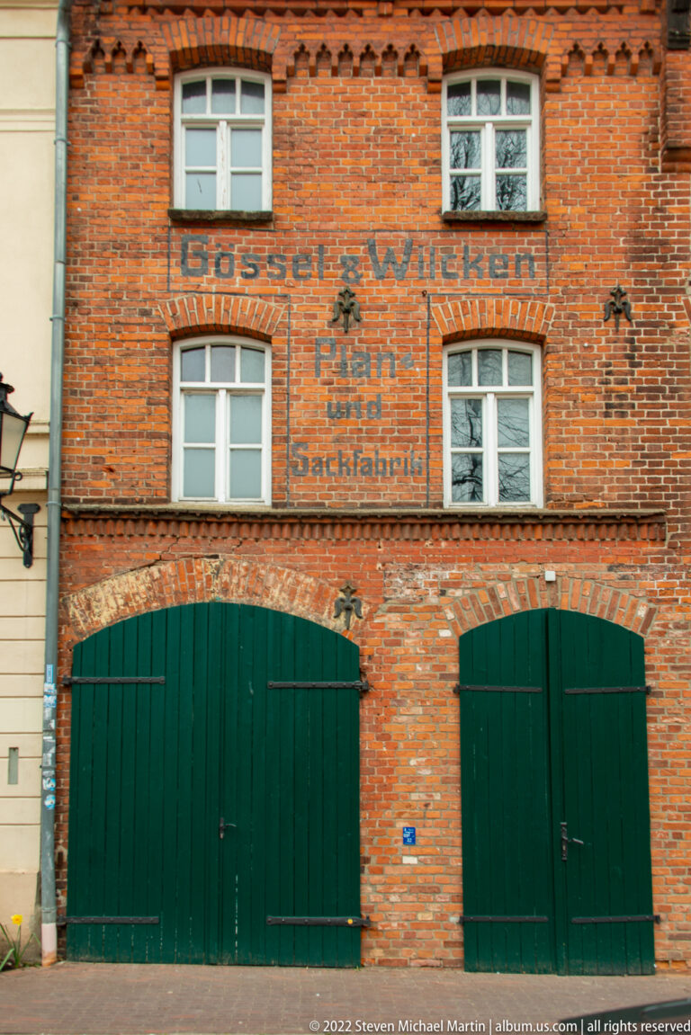 Red-brick commercial façade with faded painted German trade signage, white-framed windows, and two large green timber doors.