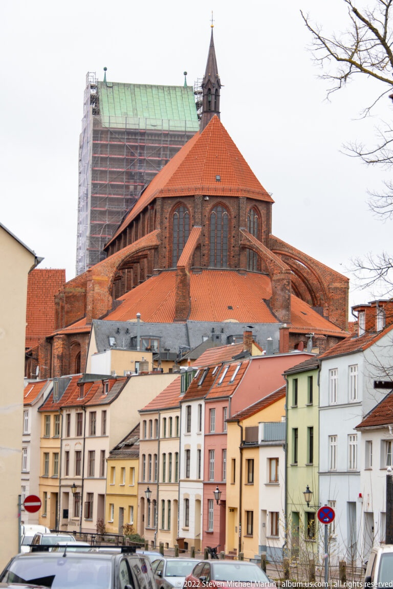 Large red-brick Gothic church with tall windows and red roofs rises behind pastel houses; part of the structure is scaffolded.
