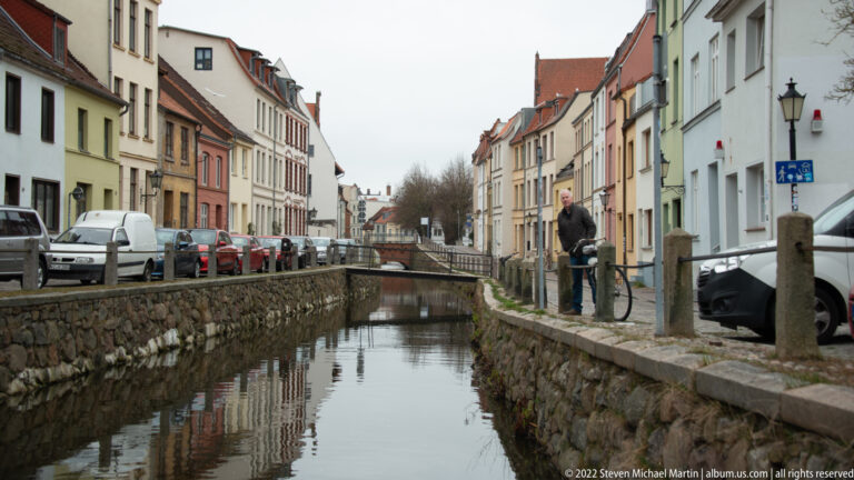 Narrow canal with stone embankments runs between historic houses, parked cars, and a pedestrian on the right bank.