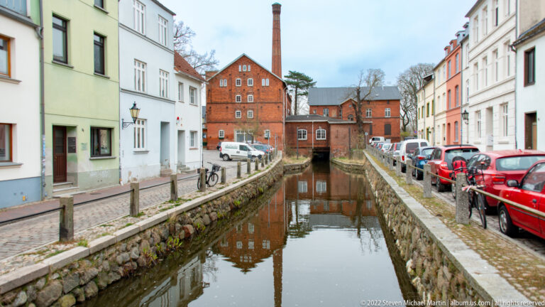 Stone-lined canal leads toward red-brick industrial buildings and a tall chimney, with houses and parked cars on both banks.
