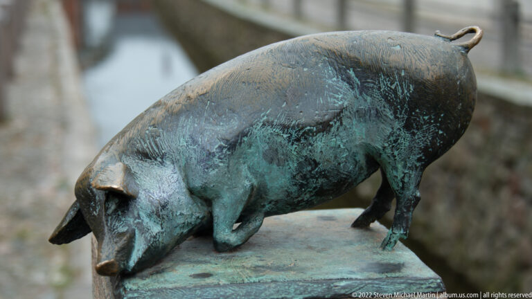 Bronze sculpture of a standing pig with lowered snout, curled tail, and mottled green patina.