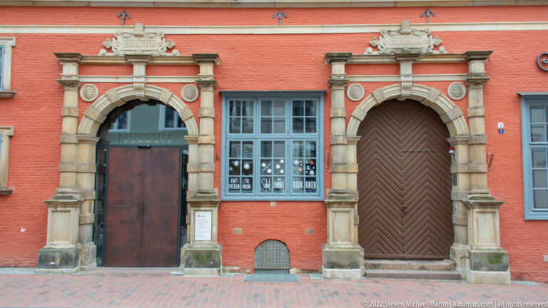 Two ornate stone-framed portals in a red façade, one with a dark door and one with a diagonal-board timber door.