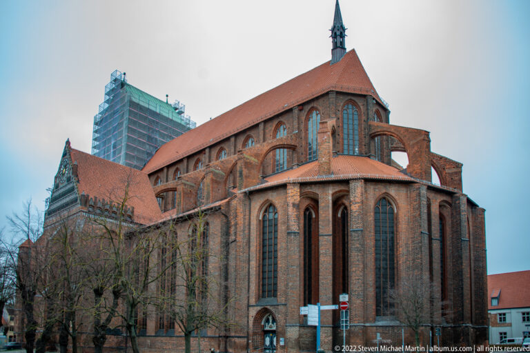 Large red-brick Gothic church with tall pointed windows, buttresses, steep tiled roof, and scaffolding around a distant tower.