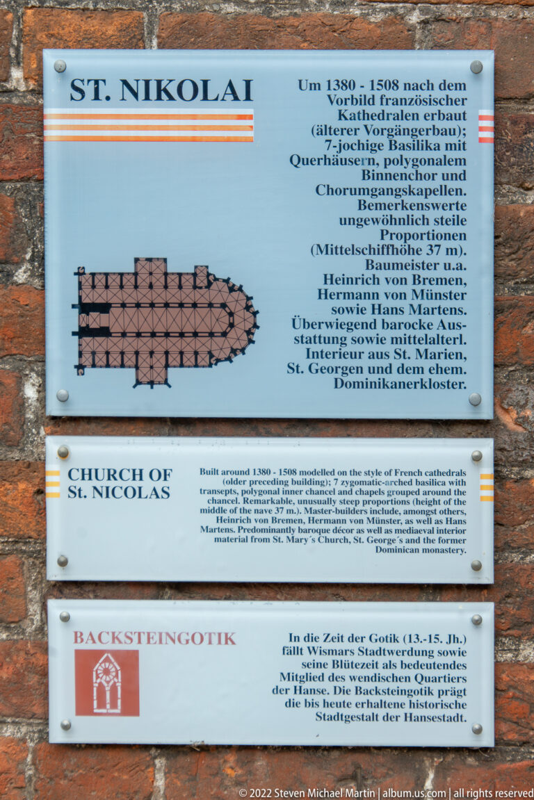 Three interpretation panels on a brick wall describe the Church of St. Nicholas and Brick Gothic in German and English.