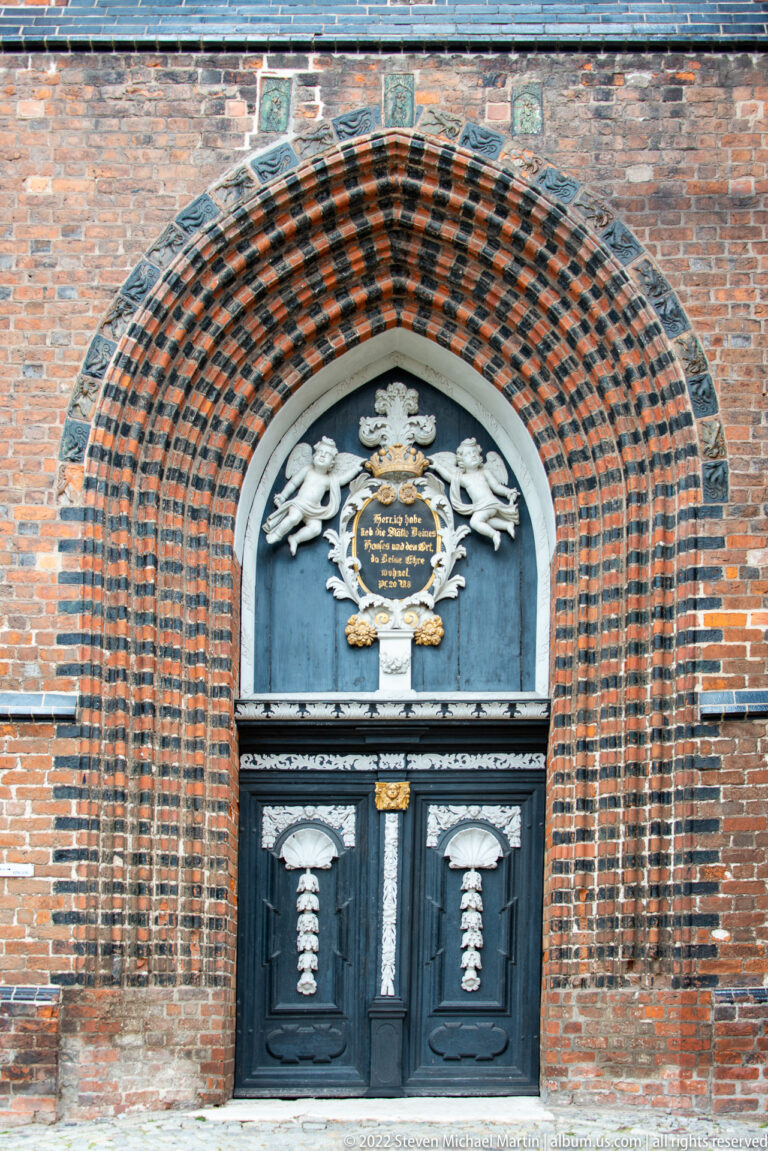 Dark double church door beneath a deep striped brick arch and an ornate heraldic cartouche with gilded lettering.