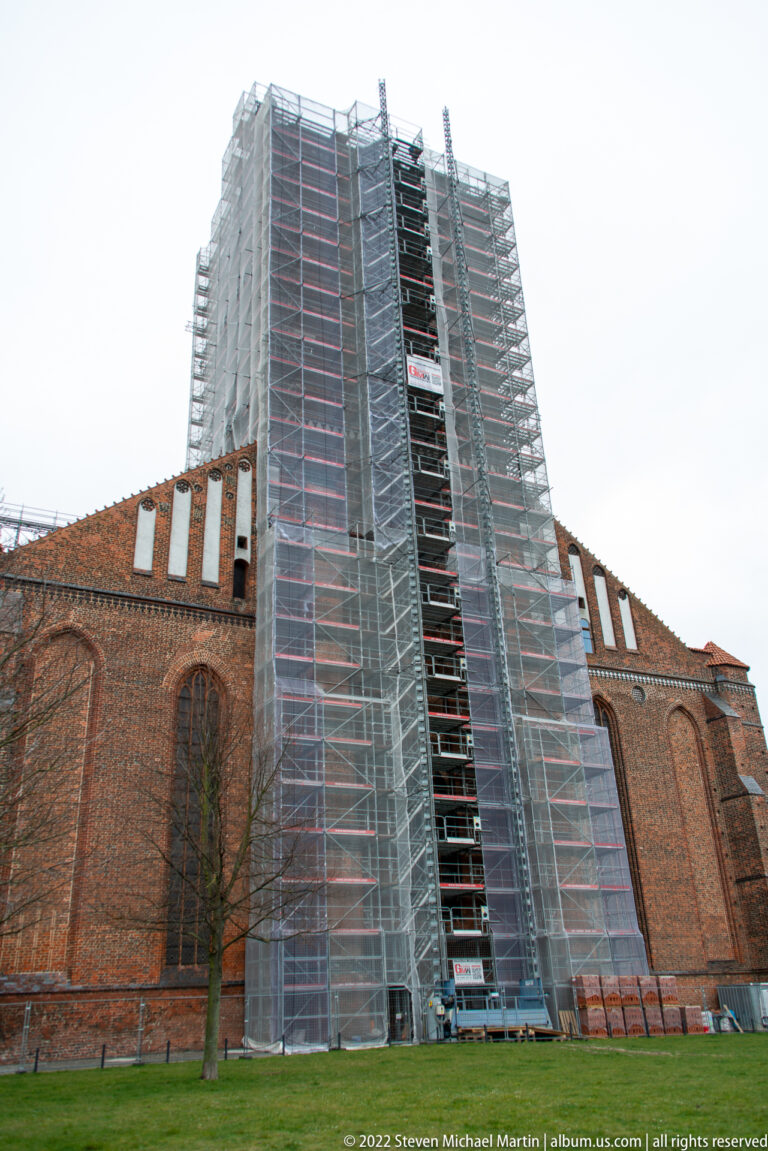Large scaffold tower covers part of a red-brick church beside a lawn, with tiled roofs and Gothic windows visible around it.