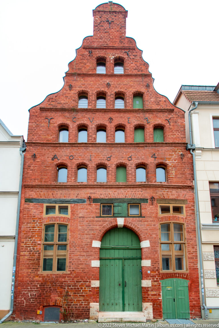 Red-brick warehouse with broad stepped gable, rows of small recessed openings, rectangular windows, and a large green central door.
