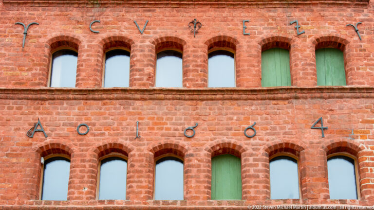 Close view of red-brick warehouse wall with two rows of small arched openings, green shutters, and large metal wall anchors.