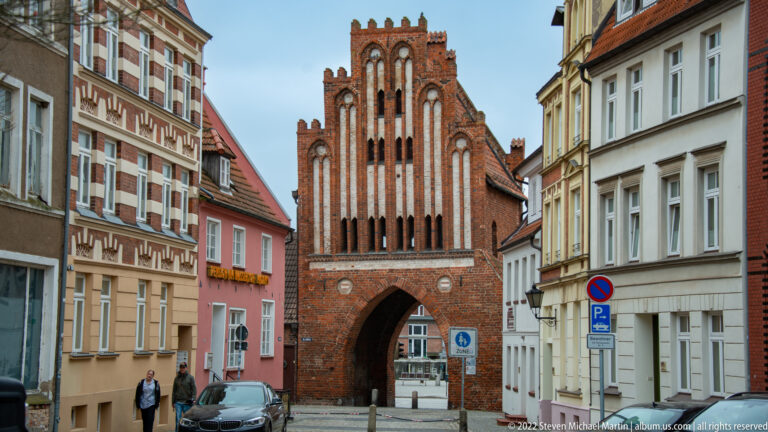 Brick Gothic city gate with pointed arch passage and tall ornate gable stands at the end of a street lined with houses.