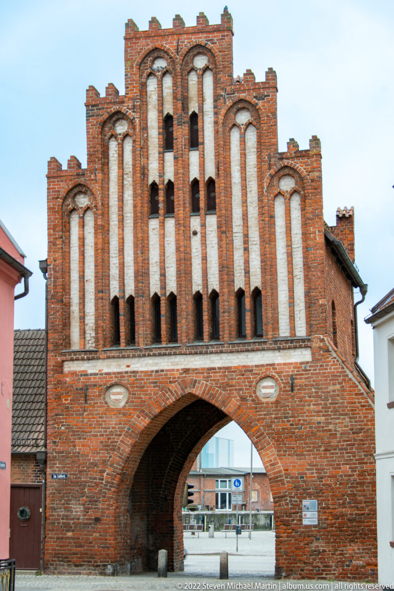 Broad brick city gate with pointed passage and symmetrical stepped gable filled with tall pale recessed panels.