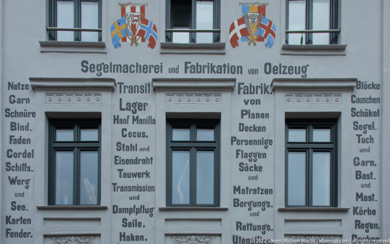 Historic German sailmaking and marine-supply advertising painted in columns around three windows on a pale façade.