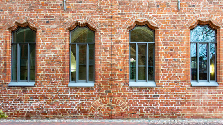 Four narrow pointed windows framed by molded red brick above a blocked arch in a historic wall.