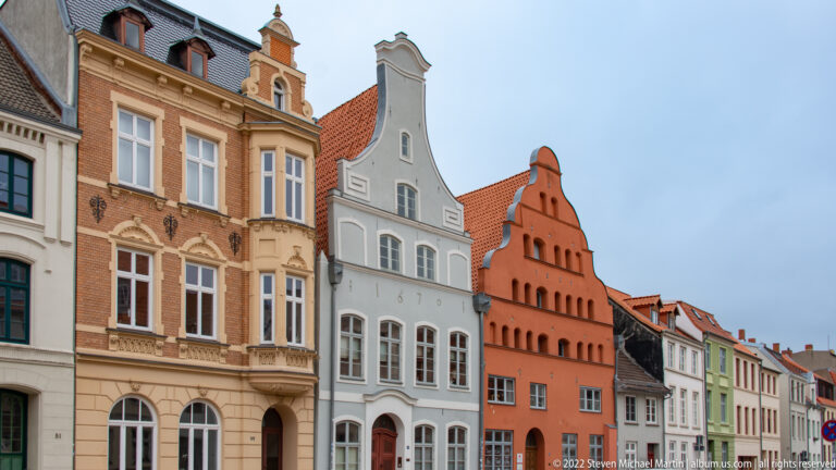 Row of historic townhouses with decorative brick, white and orange gabled façades, tall windows, and red-tiled roofs.