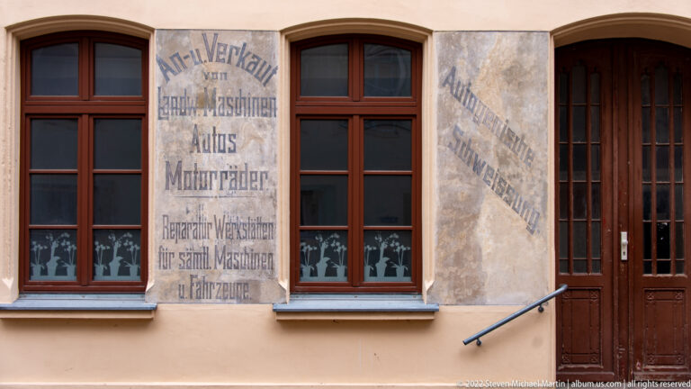Faded German commercial lettering painted on plaster panels between brown-framed windows and a tall wooden doorway.