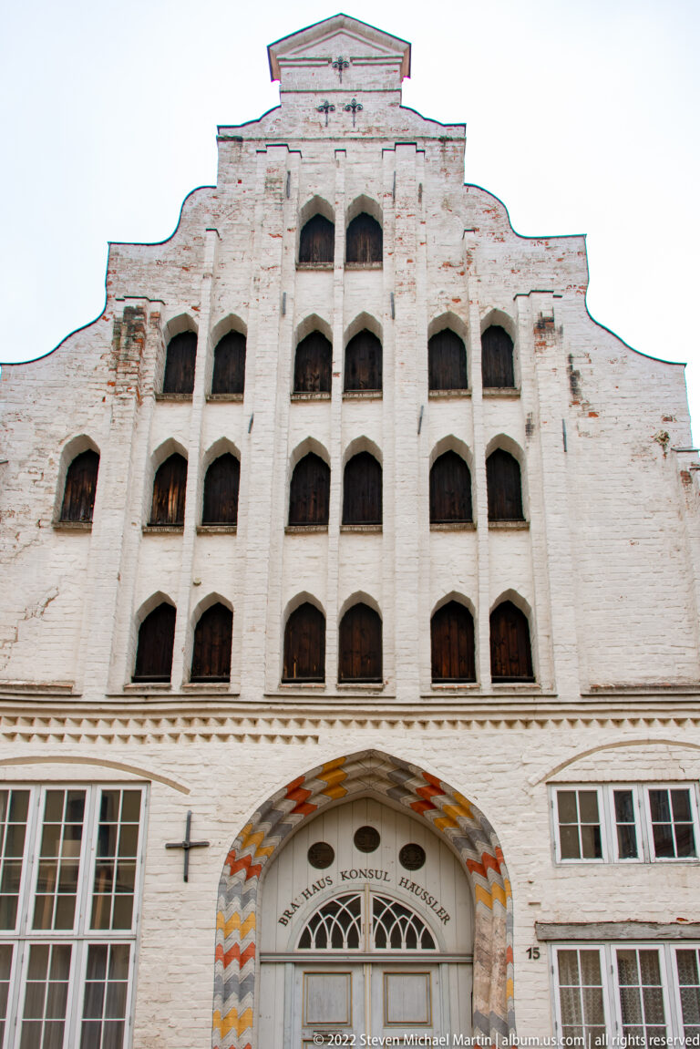 White-painted brick building with broad stepped gable, tiers of recessed openings, and a large patterned arched entrance.