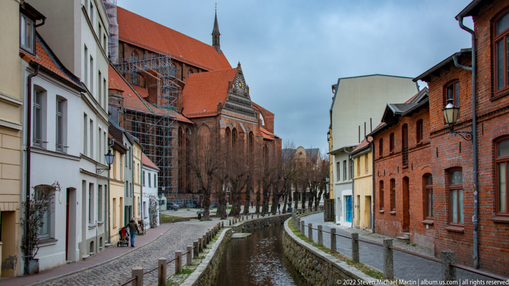 Curving stone-lined canal passes houses toward a large red-brick church with scaffolding in the background.