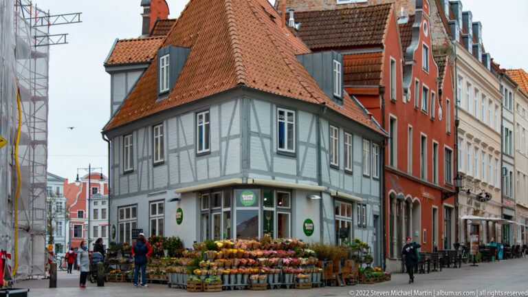 Outdoor produce display and shoppers beside a gray half-timbered corner building, with red gabled houses extending down the street.