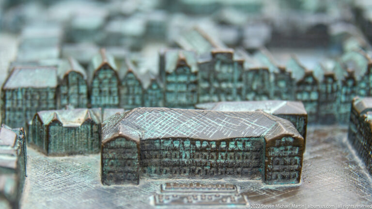 Close-up of a large rectangular building on a metal city model, with rows of miniature gabled houses blurred behind.